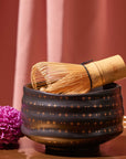 A bamboo matcha whisk rests on the Ceremonial Matcha Bowl, a handmade ceramic piece adorned with delicate 14k gold detailing and star patterns. A purple chrysanthemum flower sits nearby, while pink curtains form the background.