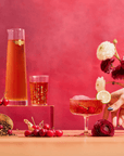 A hand holds a glass of Cherry Cola Black Tea: Ceremonial Soda Pop Tea with Sugar "Destroyer," garnished with lime, surrounded by cherries, flowers, a carafe, and another glass on wood against a vibrant pink-red backdrop.