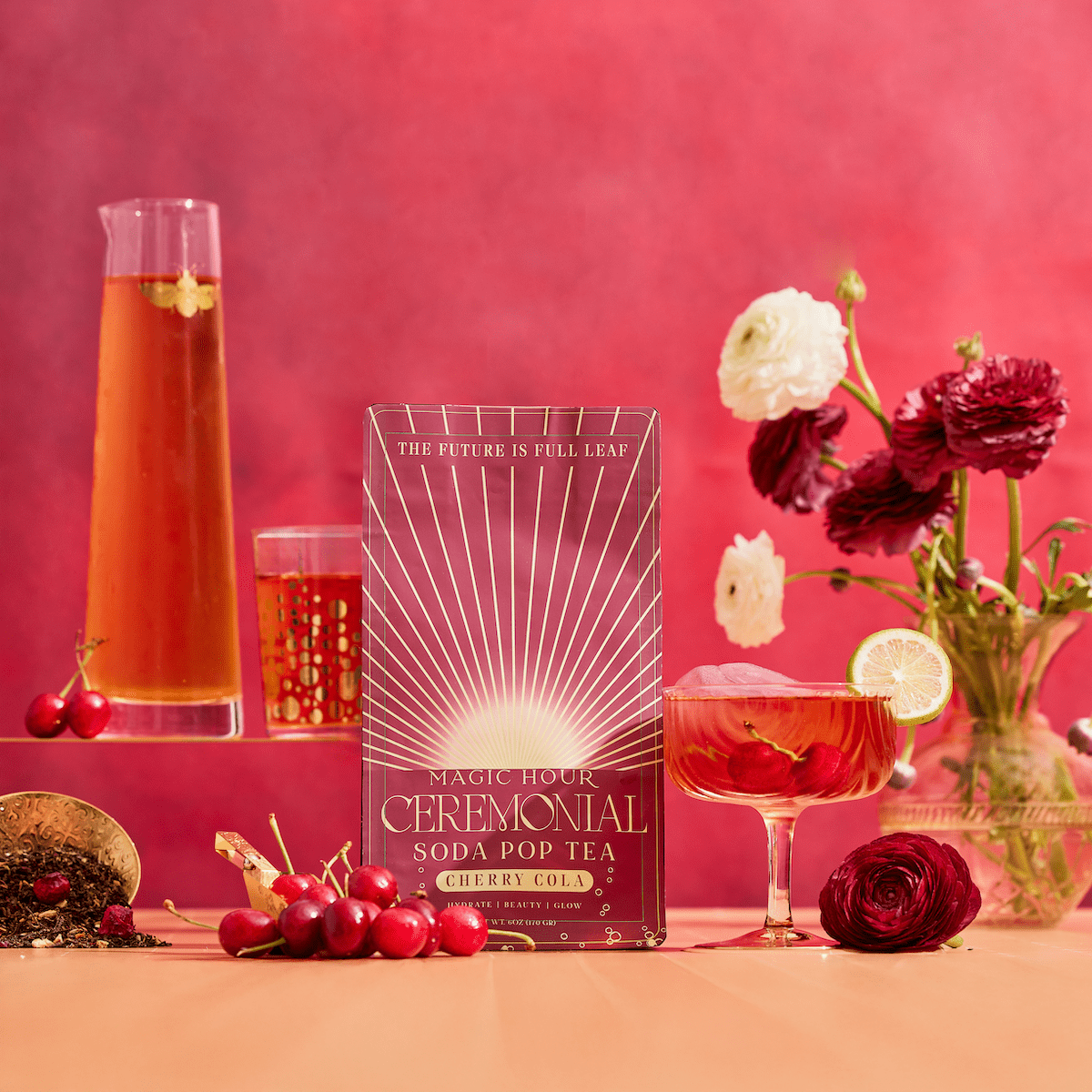 A vibrant display features a Cherry Cola Black Tea: Ceremonial Soda Pop Tea with Sugar "Destroyer" box, bottle and glass of cherry tea, fresh cherries, Assam black tea leaves, and red and white flowers on a pink-red background.