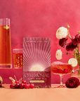 A vibrant display features a Cherry Cola Black Tea: Ceremonial Soda Pop Tea with Sugar "Destroyer" box, bottle and glass of cherry tea, fresh cherries, Assam black tea leaves, and red and white flowers on a pink-red background.