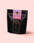 A black resealable pouch with gold star speckles labeled "Child’s Pose™ Herbal Adaptogen Tea Blend for Sleep & Restful Calm" stands upright against a light pink background.