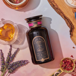 A dark glass jar labeled "Child’s Pose™ Herbal Adaptogen Tea Blend for Sleep & Restful Calm" sits on a pink and beige background, surrounded by a heart-shaped cup of herbal tea, dried rose petals, lavender sprigs, and a golden teaspoon.