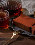 A glass plate holds rectangular slices of Small Batch Magic Valentine’s 2026 chocolate cake dusted with cocoa, beside a vintage fork, chocolate cake tea, and a glass pitcher on a wooden table with a gray cloth background.