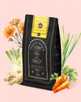A black and gold "Citrine Cleanse™ Herbal Tea" package rests among carrots, organic Hawaiian ginger, fennel, calendula flowers, and lemongrass—showcasing a caffeine-free detox elixir on a pale pink background.