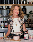 A smiling woman stands behind a counter adorned with beauty products, a bowl, and a whisk—evoking the inviting atmosphere of The Soul of Tea: A Three Day Retreat in Ojai, CA. Shelves lined with bottles and a glowing circular sign create an ambiance that seamlessly blends the sophistication of skincare with the serene rituals celebrated during this immersive Ojai tea retreat.