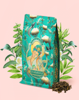 A teal floral tea bag labeled "Compassion: Quan Yin Original Style Oolong," adorned with an illustration of a woman amid green leaves and white flowers, is displayed beside a small pile of loose oolong tea on a light pink background.