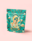 A teal and gold pouch labeled "Compassion: Quan Yin Original Style Oolong" displays a serene woman amid flowers, birds, and leaves on a soft pink background, capturing the calming essence of this unique Oolong tea.