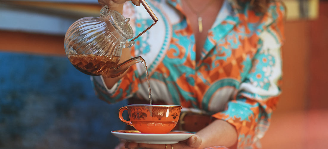 A person wearing a colorful, patterned shirt pours tea from a glass teapot into an orange floral teacup. The person holds the saucer with one hand while carefully pouring with the other.