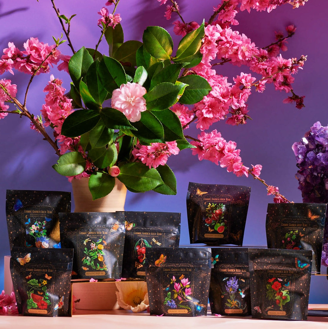 A variety of tea packages, including the Cosmic Garden Iced Tea Sampler Set, are arranged around a potted plant with pink flowers on a wooden table. The packages feature vibrant illustrations set against a blue-to-purple gradient backdrop, enhanced with a crystal cluster for added decor.