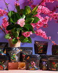 A variety of tea packages, including the Cosmic Garden Iced Tea Sampler Set, are arranged around a potted plant with pink flowers on a wooden table. The packages feature vibrant illustrations set against a blue-to-purple gradient backdrop, enhanced with a crystal cluster for added decor.