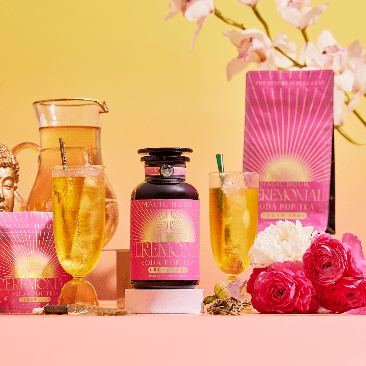 A vibrant display of Cream Soda White Tea: Ceremonial Soda Tea with Sugar Destroyer, in pink and gold packaging, alongside two iced tea glasses, a pitcher, flowers, and a Buddha statue on a bright pink-yellow gradient background.