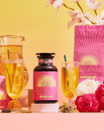 A vibrant display of Cream Soda White Tea: Ceremonial Soda Tea with Sugar Destroyer, in pink and gold packaging, alongside two iced tea glasses, a pitcher, flowers, and a Buddha statue on a bright pink-yellow gradient background.