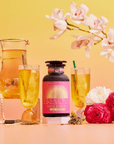 A bottle of Cream Soda White Tea: Ceremonial Soda Tea with Sugar Destroyer is displayed with two glasses of iced tea, a pitcher, pink flowers, a golden Buddha statue, pearls, and tea leaves on a yellow-to-pink gradient background with orchids.