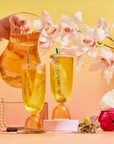 A hand pours golden Cream Soda White Tea: Ceremonial Soda Tea with Sugar Destroyer into two tall glasses, surrounded by pink orchids, a gold Buddha statue, pearls, and vibrant flowers on a pastel surface.