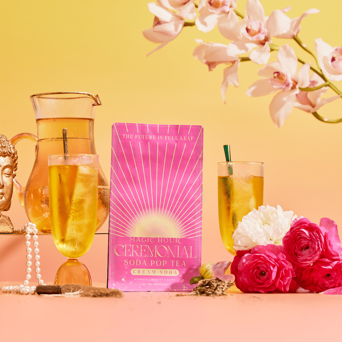 A vibrant scene featuring a pink bag of Cream Soda White Tea: Ceremonial Soda Tea with Sugar Destroyer, two glasses of iced tea, a glass pitcher, pink and white flowers, a gold Buddha head, pearls, and orchids on a warm gradient background.