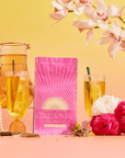 A vibrant scene featuring a pink bag of Cream Soda White Tea: Ceremonial Soda Tea with Sugar Destroyer, two glasses of iced tea, a glass pitcher, pink and white flowers, a gold Buddha head, pearls, and orchids on a warm gradient background.