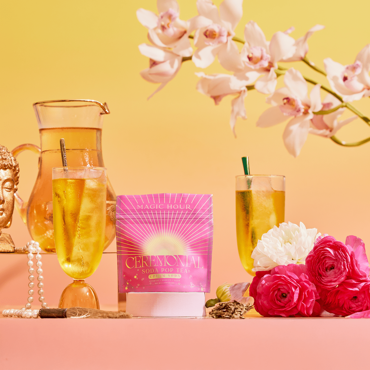A vibrant display showcases a pink Cream Soda White Tea: Ceremonial Soda Tea with Sugar Destroyer box, two iced tea glasses, a pitcher, pink and white flowers, pearls, a Buddha statue, and orchids on a yellow-to-pink gradient background.