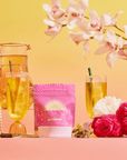 A vibrant display showcases a pink Cream Soda White Tea: Ceremonial Soda Tea with Sugar Destroyer box, two iced tea glasses, a pitcher, pink and white flowers, pearls, a Buddha statue, and orchids on a yellow-to-pink gradient background.