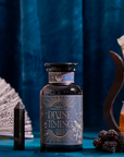 A bottle of Divine Timing Balance rests on a blue table alongside a crystal, dried fruit, blackberries, a fan, and a teapot with tea, set before a luxurious blue velvet curtain.