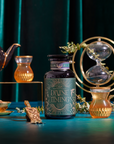 A jar labeled "Divine Timing Calm" sits on a teal table, surrounded by glass cups of tea, a teapot, an hourglass, and tea leaves, all set against a rich green velvet backdrop.