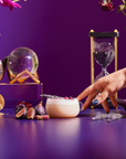 A hand touches a glass of Divine Timing Rest, a creamy cocktail garnished with flowers and lavender, surrounded by figs, petals, a golden tea set, an hourglass, and vibrant purple blooms on a purple surface and background.