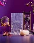 A styled scene features the "Divine Timing Rest" card deck at the center, surrounded by an hourglass, crystal ball, figs, a latte, flowers, and crystals on a purple background.