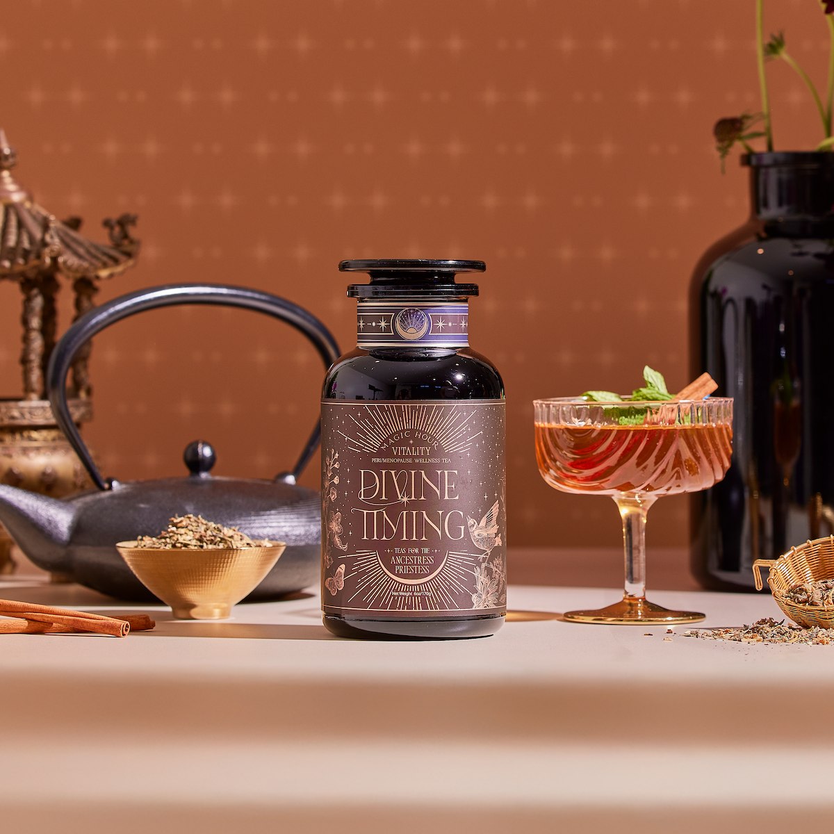 A jar of Divine Timing Vitality rests on a table next to a glass of herbal drink, teapot, cinnamon sticks, loose herbs, and dark vases, all set against a warm patterned backdrop.