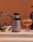 A jar of Divine Timing Vitality rests on a table next to a glass of herbal drink, teapot, cinnamon sticks, loose herbs, and dark vases, all set against a warm patterned backdrop.