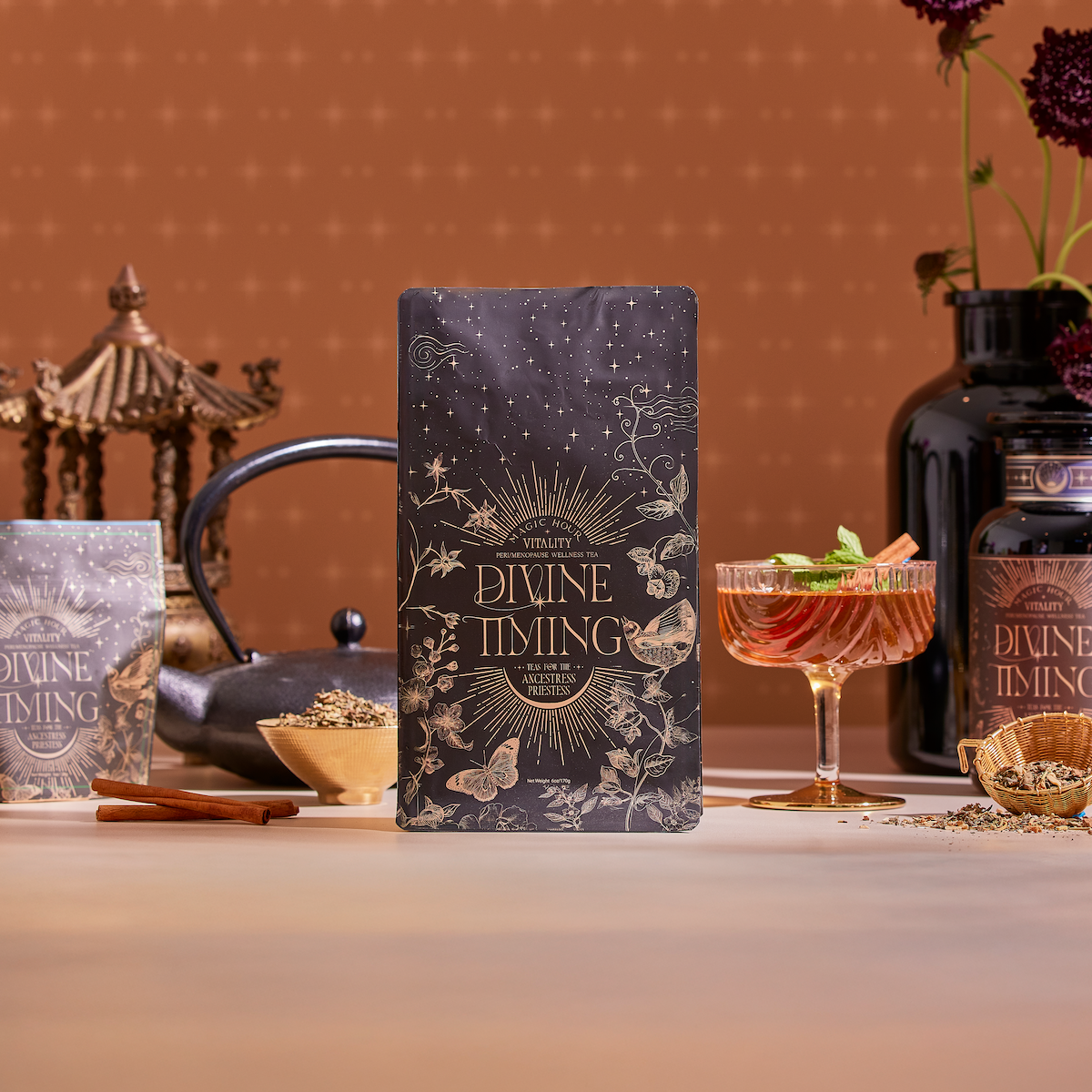 A decorative tea package labeled "Divine Timing Vitality" sits on a table with loose tea, a teapot, iced tea, and jars, set against a warm floral background with a pagoda—crafted to support vitality and adaptogenic benefits.