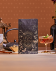 A decorative tea package labeled "Divine Timing Vitality" sits on a table with loose tea, a teapot, iced tea, and jars, set against a warm floral background with a pagoda—crafted to support vitality and adaptogenic benefits.