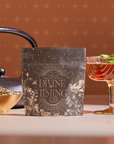 A packet labeled “Divine Timing Vitality” herbal tea sits on a table with loose herbs, a black teapot, and a glass of mint-garnished tea—showcasing its adaptogenic and metabolism-supporting herbs in warm light.