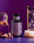 A bottle labeled “Divine Timing Rest” rests on a purple surface with figs, flowers, a latte, hourglass, and glass orb nearby. The purple gradient background adds a mystical atmosphere.