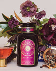 A jar of Queen of the Tropics: Rosé all Day Alternative Tea is shown with a pink tea cocktail, a tea strainer with loose leaves, and dark purple flowers—a perfect tropical fruit blend and refreshing rosé wine alternative.