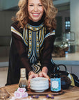 A smiling woman with wavy hair sits at a table with a tea set, featuring the Ceremonial Matcha Bowl—handmade with 14k gold detailing—alongside a jar labeled “cultivate,” a matcha whisk, dried flowers, and decorative items arranged on a wooden tray in a modern kitchen. The scene highlights traditional artisan craftsmanship and warm contemporary style.