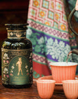 A person in colorful patterned clothing pours hot water into a cup beside a "Freya The Radiant: Jasmine Bai Hao Oolong with Bergamot & Berry" tea jar, with more cups and an ornate teapot on a wooden table.