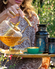 Outdoors among greenery and flowers, someone in patterned clothing pours Hathor of Joy: Honey Nectar Herbal Tea from a glass teapot into a green teacup on a tray, with two ornate "Magic Hour" containers beside the cup.