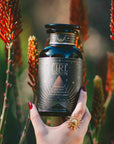 A hand with red nails and a sun-shaped ring holds a dark bottle labeled “Fire Ignition” amid tall orange flowers and green stems in soft sunlight, evoking the energizing blend of Fire: Spiced Black Tea with Ginger and Cardamom to Ignite Courage and Vitality.