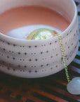 A creamy pink drink fills a blush cup adorned with gold stars, while the Rose Quartz Heart Tea Strainer—with its delicate chain and heart-shaped charm—rests gracefully inside. This elegant tool is perfect for elevating your tea ritual, as the cup sits atop a patterned surface, inviting tranquil moments inspired by rose quartz serenity.