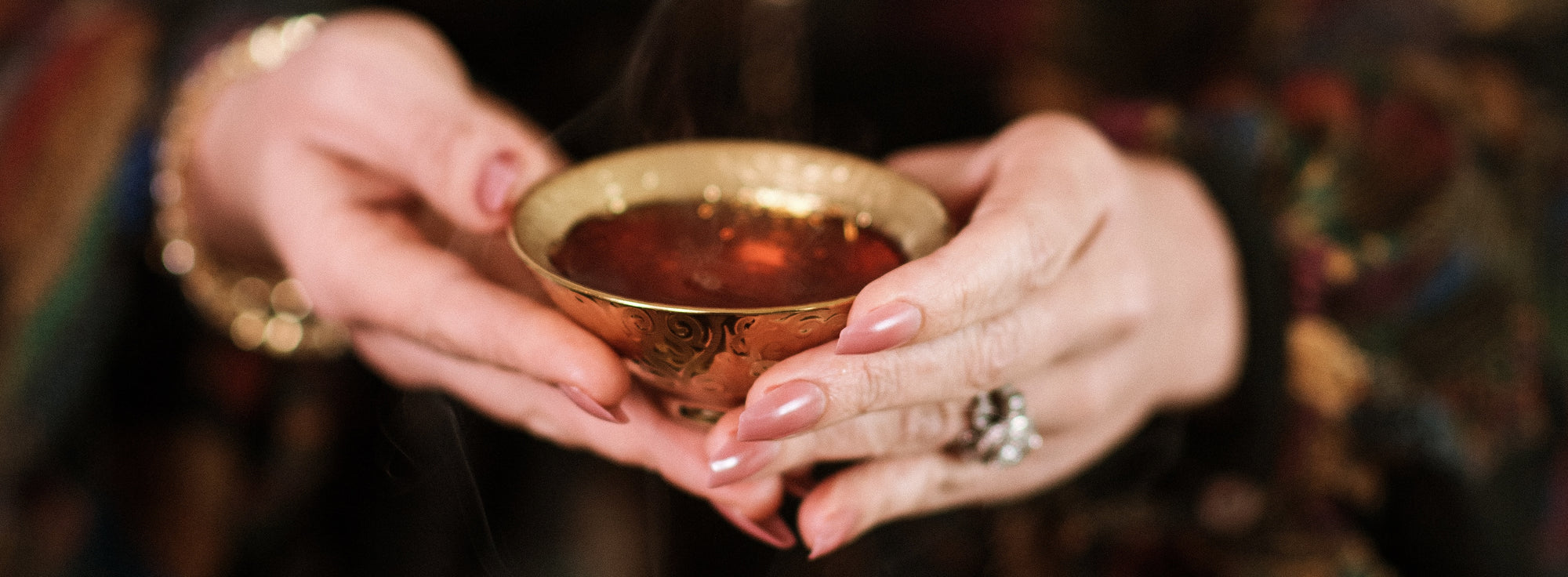 Close-up of a person with manicured nails holding a small, ornate brass bowl filled with a dark liquid. The person's hands and part of a colorful, patterned garment are visible, with a ring on one finger.