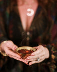 A person with painted nails and a crescent moon necklace holds a small brass bowl of dark tea—possibly brewed from the Tea Essentials + In Person & Virtual Tea School Course—complementing their colorful, patterned outfit.