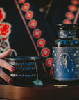 A person with painted nails and a turquoise ring holds a black mug with star patterns. Beside it sits a "Skadi The Sovereign" Magic Hour tea jar, containing a ceremonial berry-infused puerh tea for winter resilience on wood.
