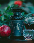 A black jar labeled "Monthly Magic Tea Subscription Box" stands by a whole pomegranate, as a glass teapot pours red tea into a cup—inviting you to a ceremonial tea experience outdoors among lush green foliage.