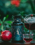 A glass teapot pours hibiscus tea into a small cup beside a pomegranate and a floral-labeled "Persephone's Potion: Pomegranate & Jasmine Green Tea" jar on wood, with blurred green foliage in the background.