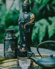 A tranquil arrangement showcases a black statue holding a scroll, accompanied by a black jar adorned with intricate designs, and a lit black candle placed in front of the statue. Next to them sits The Oracle: Lemon Lavender Tea for Supporting the Gut-Brain Axis Case Packs from Magic Hour, steeped in a cast iron teapot and poured into a small glass cup on a golden tray. The serene setting is complemented by greenery in the background, evoking an outdoor oasis.