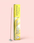 A yellow box labeled “Diamond Incense” stands upright, its fragrance promising a calming ambiance, next to a single incense stick held in a small white holder, set against a pale pink background.