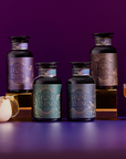 Four elegant jars from the Divine Timing Tea Bundle for menopause support are displayed on gold risers against a purple background, accompanied by a berry milk drink, a coupe glass cocktail, and a deep purple flower arrangement.
