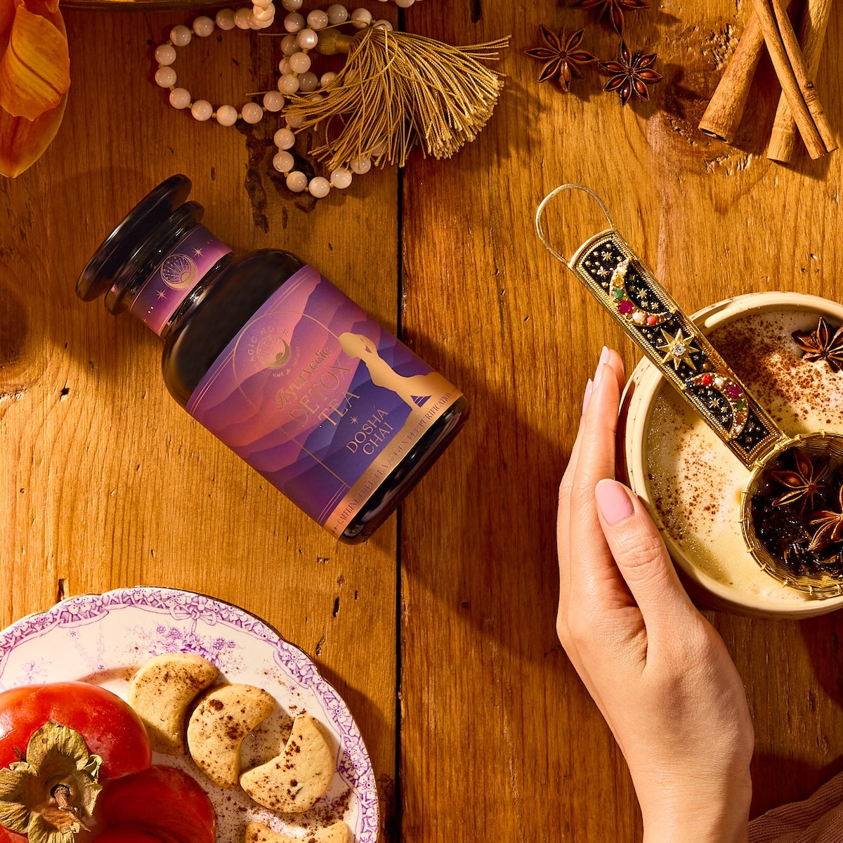 A hand holds a bowl of spiced milk next to a bottle labeled “Dosha Herbal” on a wooden table with cookies, persimmon slices, a beaded necklace, and spices such as cinnamon sticks and star anise.