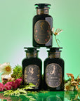 Three dark apothecary jars, labeled “Virgo,” “Capricorn,” and “Dawn,” rest among ferns, flowers, and mushrooms on a green backdrop, creating a mystical vibe—an ideal addition to the Earth Sign Astrology Bundle.