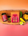 A wooden board displays a jar and pouch of Teasoning: Chili Fruit Spice tea, surrounded by loose tea, matcha hibiscus, jalapeño, yellow chile, pineapple chunks, halved lime, and tropical seasoning on an orange-to-pink gradient backdrop.