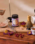 A tea-making scene features the Flower Powder: Matcha Bundle on a wooden board with matcha powder, dried flowers, a bamboo whisk, teapot, cup of tea, and incense. Matcha from the bundle is being sifted into a glass bowl.